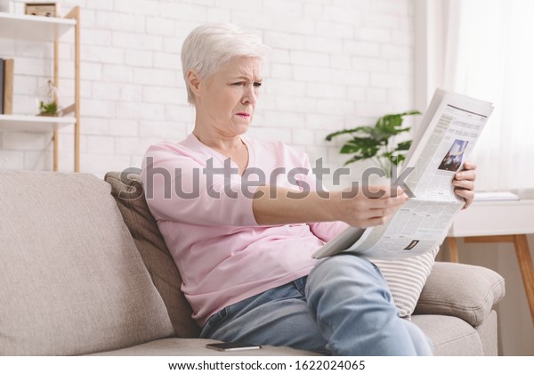 Bad sight problem. Senior lady\
squinting and holding newspaper far from eyes at home, free\
space