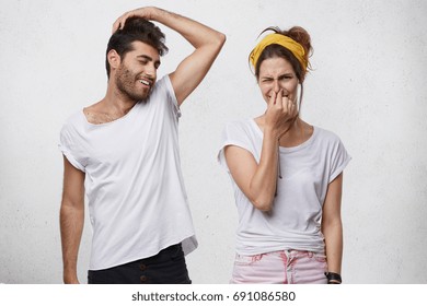 Bad smell and body odor. Picture of fastidious young woman pinching her nose disgusted with terrible sweat, coming out from bearded man's armpit who is standing next to her, raising his arm