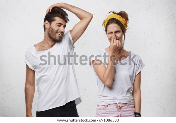 Bad\
smell and body odor. Picture of fastidious young woman pinching her\
nose disgusted with terrible sweat, coming out from bearded man\'s\
armpit who is standing next to her, raising his\
arm