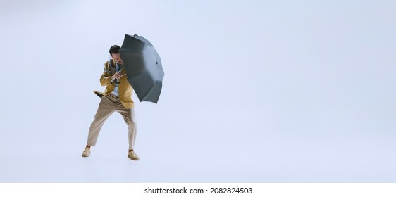 Bad Weather. Portrait Of Young Man Dressed In 50s, 60s Style Fights The Wind With Umbrella Isolated On White Background. Autumn Fashion Collection. Concept Of Culture, Art, Wellbeing, Beauty And Ad.