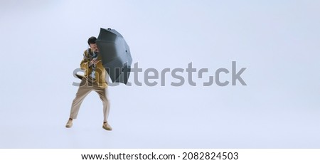 Bad weather. Portrait of young man dressed in 50s, 60s style fights the wind with umbrella isolated on white background. Autumn fashion collection. Concept of culture, art, wellbeing, beauty and ad.