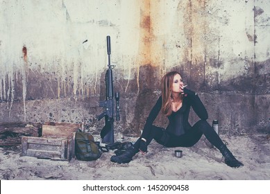 A badass lady in a black catsuit smokes a cigarette while sitting near her combat equipment.