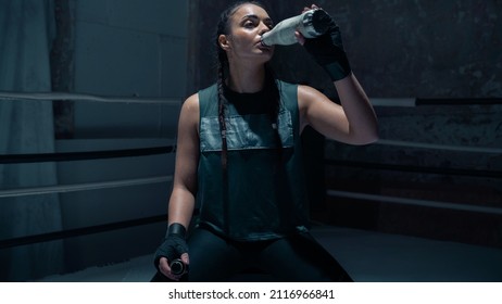 Badass Looking Ethnic Female Kick Boxer With Braids Kneeling on the Mat in The Middle of a Boxing Ring Drinking Water In Between Sparring Sessions and Training With Her Coach. Confident Woman Athlete. - Powered by Shutterstock