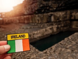 Badge With Sign Ireland And Irish National Flag In Focus, The Worm Hole Out Of Focus In The Background. Sun Flare. Aran Island, County Galway. Travel And Tourism Theme. Explore Irish Landmark Concept