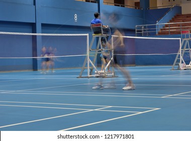 A Badminton Court Number 8 In Action Seeing Movements Of Double Female Players Playing While Focusing On An Umpire On Duty In The Mid Court.