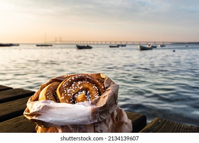 Bag Of Cinnamon Rolls Or Buns On A Wooden Boardwalk By The Ocean At Dusk With The Oresund Bridge. Cosy Swedish Fika Break By The Baltic Sea With Cinnamon Bun At Sunset, Typical Scandinavian Pastry