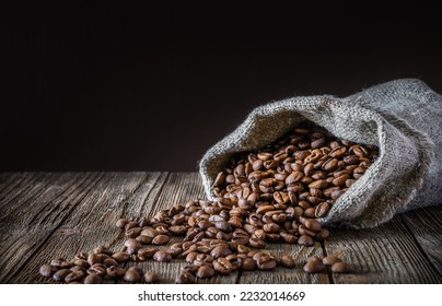 A bag of coarse canvas of coffee beans is lying on its side. Coffee beans scattered on the wooden tabletop.  Background with dark wood texture. - Shutterstock ID 2232014669