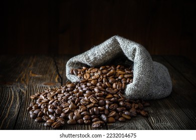 A bag of coarse canvas of coffee beans is lying on its side. Coffee beans scattered on the wooden tabletop.  Background with dark wood texture. - Shutterstock ID 2232014675