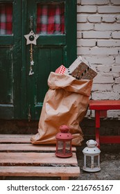 Bag Of Gifts At Christmas Under The Door. Delivery Of New Year's Gifts. Santa Claus Bag With Congratulations. Xmas Porch Decor, Kraft Paper Bag With Christmas Boxes