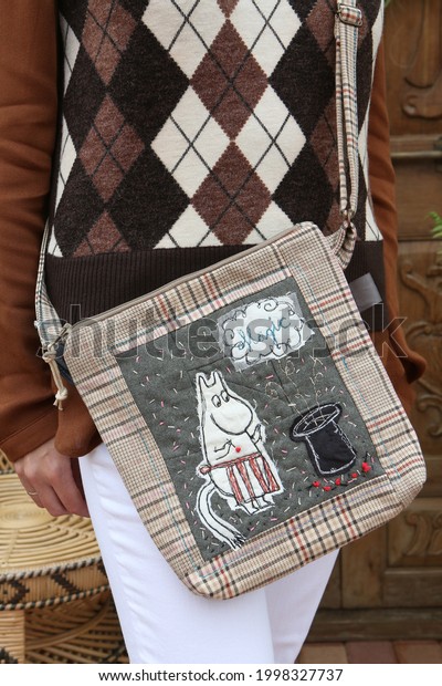 Bag. Look of russian fashion woman. Spring outfit. Girl with stylish bag, beige coat, white jeans, grey sweater. Bag, purse with embroidery with Mummy trolls. Street style, combination of clothes