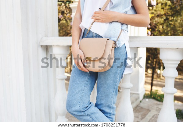 Bag in women\'s hands. Details of casual summer or\
spring outfit. Woman wearing blue jeans, white t shirt, small beige\
cross body bag standing outdoors. Everyday look. Street fashion. No\
face.