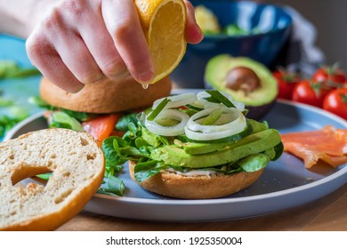 Bagel sandwiches in the making. Gluten free toasted bagel with avocado, mayo, lettuce, cucumber and onion. Hand squeezes lemon on top and salmon bagel background. Healthy breakfast, fun vibrant colors - Powered by Shutterstock