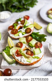 Bagels sandwich topped with cream cheese, farm cottage cheese, avocado, tomatoes, cucumbers and salad leaves, grey tilled background. Healthy breakfast food.