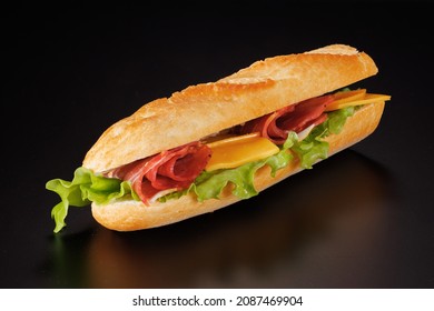 Baguette Sandwich With Ham, Smoked Sausage, Cheese, And Green Salad On A Black Background.