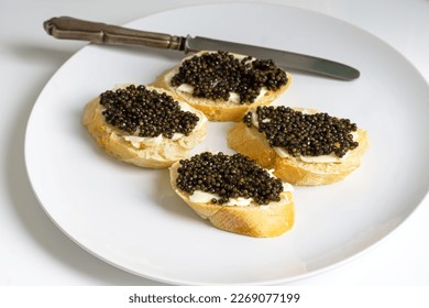 Baguette slices with black caviar and butter on a white plate, silver knife aside. Natural sturgeon luxury delicacy sandwich closeup. Seafood delicatessen and bread appetizer. - Shutterstock ID 2269077199