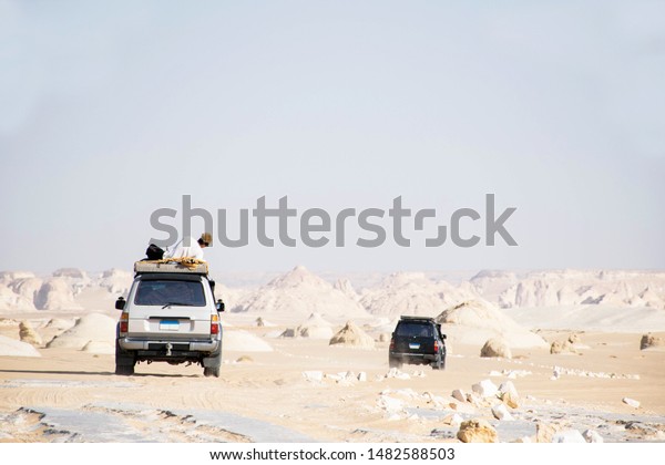 Bahariya, Egypt - 06-07-2019: Two cars driving in\
the white desert with man on the roof enjoying the view. Exploring\
the wilderness. Scenic sahara landscape egypt. White rock\
formations. Travel\
scene.