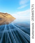 Baikal Lake on cold winter day. Beautiful landscape with blue clear ice with cracks and coastal hills in Kharyuzovaya Bay. Ice travels and winter hiking. Natural blue background, ice texture