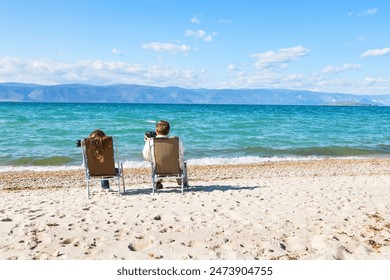 Baikal Lake in summer. Two tourists sit on sandy beach of Olkhon Island, look at the mountains through binoculars, take pictures of the Small Sea and enjoy outdoor recreation. Summer travel and rest - Powered by Shutterstock