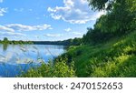 A bait boxes, a bucket with lure, a feeder rods for bream fishing mounted on stands next to bushes on the high grassy river bank. On the far bank is a forest. Sunny summer weather and blue sky