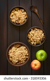 Baked Apple Crumble Or Crisp In Rustic Bowls, Wooden Spoons And Fresh Apples On The Side, Photographed Overhead On Dark Wood With Natural Light