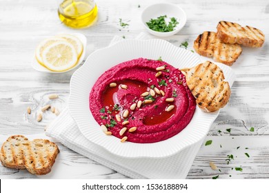 Baked Beet Hummus with toasted bread in a white ceramic plate on a white wooden background. Top view