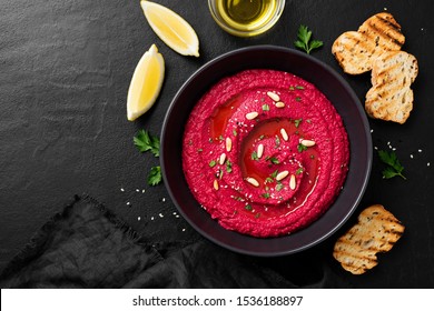 Baked Beet Hummus with toasted bread  in a black ceramic bowl on a dark background. Top view