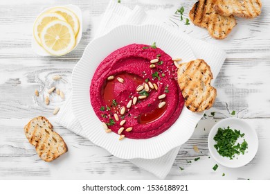 Baked Beet Hummus with toasted bread in a white ceramic plate on a white wooden background. Top view