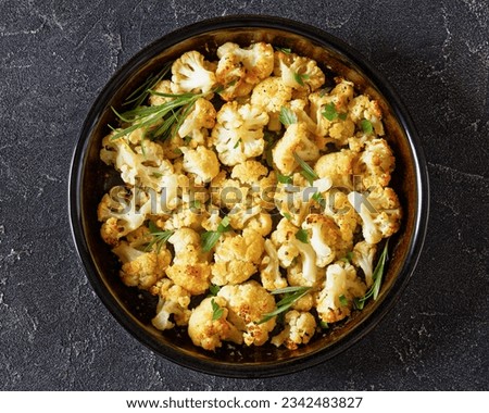 baked cauliflower florets dredged with grated parmesan and herbs in baking dish on concrete table, horizontal view from above, flat lay, close-up