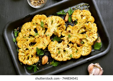 Baked cauliflower steaks with herbs and spices on baking sheet over black stone background.