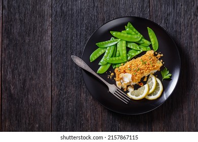 Baked Cod with Crackers butter toppings served with boiled snow peas and lemon on black plate on dark wooden table, horizontal view from above, flat lay, free space, close-up