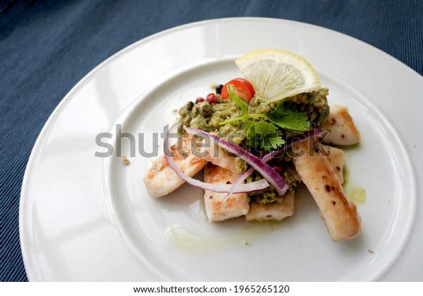 Baked Cod fish\
with green sauce (salsa verde) with lemon, red onion, coriander,\
red chili, pickles and capers. Basque cuisine. On white plate. On\
the blue background.\
Close-up.
