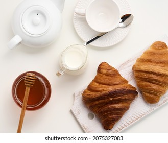 Baked Croissants, White Ceramic Teapot And Empty Cup And Saucer, Jar Of Honey On A White Table, Top View. Breakfast