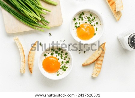 Baked eggs (Eggs en cocotte) with cream, сheese and green onion in portion ceramic bowl and toasted bread slices over white background. Comfort food for breakfast. Top view, flat lay