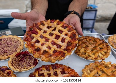 Baked goods at outdoor agriculture fair. A market trader is seen close up, holding a freshly baked fruit tart with pastry topping by a display of freshly baked goods during a farmer's market.