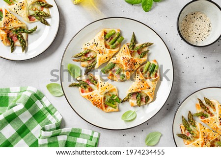 Baked green asparagus with ham and cheese in puff pastry sprinkled with sesame seeds and green basil leaves. Light gray stone background. top view