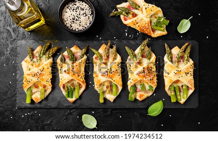 Baked green asparagus with ham and cheese in puff pastry sprinkled with sesame seeds and green basil leaves. Black stone background, top view