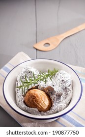 Baked Jacket Potato In Aluminium Foil Hot Off The Barbecue Lying On The Crumpled Foil Wrapper In A Basin With Fresh Rosemary, High Angle View With Copyspace