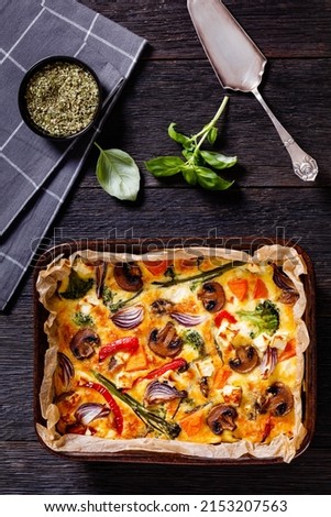 baked in oven vegetable frittata of beaten eggs, red onion, pumpkin or batata, sweet potato, yam, broccoli, red pepper and mushrooms in a baking dish on dark wood table, flat lay, vertical view