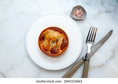 Baked Pie With Pastry Crust On A White Ceramic Plate, Irregular Shaped Grey Small Saucer Of Salt And Cutlery From An Overhead View  On White Marble Texture Kitchen Counter.
