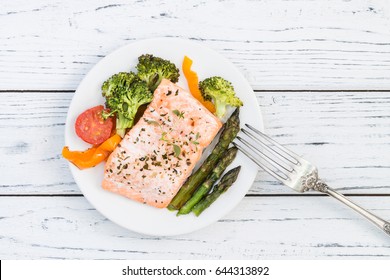 Baked salmon fillet with pepper, red cherry tomatoes, asparagus, broccoli and fresh thyme served on white plate. Wooden table. Lunch or dinner concept. Top view