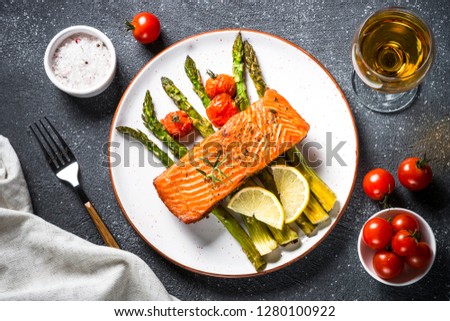 Baked salmon fish fillet with asparagus and tomato with glass wine on black stone table. Top view.