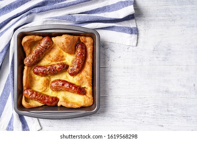 Baked Yorkshire Pudding With English Roasted Sausages Served On A Square Cake Pan On A White Wooden Table, Copy Space, Horizontal View