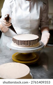 A baker is adding chocolate icing to a round cake in a kitchen. The baker uses a piping bag to create a design on the cake. The workspace is tidy with cake forms visible.