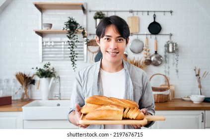 Baker Asian man smiling holds in his hands a tray with croissants baked in the bakery. Handsome baker in uniform holding baguettes with bread shelves on the background at kitchen homemade. - Powered by Shutterstock