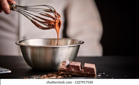 Baker or chef whisking melted chocolate in a bowl with an old metal wire whisk in a close up on his hand and utensils - Shutterstock ID 1327917530