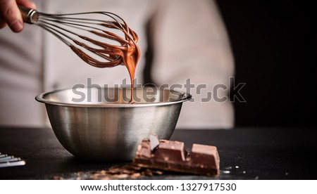 Baker or chef whisking melted chocolate in a bowl with an old metal wire whisk in a close up on his hand and utensils