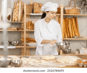 Baker girl in white uniform and chefs hat with bowl and whisk in her hands. She whips dough, combines and mixes ingredients, creates dough for sweet pastries - Powered by Shutterstock