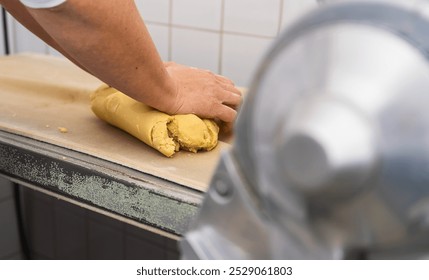 baker kneading a block of cookie dough on a countertop in a bakery, preparing it for further processing, with a rolling machine visible in the background - Powered by Shutterstock