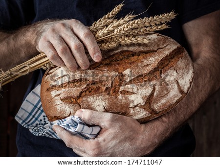 Baker man holding rustic organic loaf of  bread and wheat in hands - rural bakery. Natural light, moody still life.
