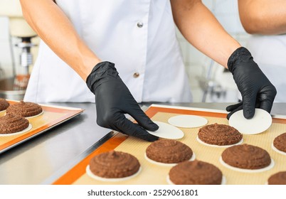 baker wearing black gloves is carefully placing wafers under freshly piped gingerbread dough on a baking sheet, preparing the cookies for baking in a commercial kitchen - Powered by Shutterstock